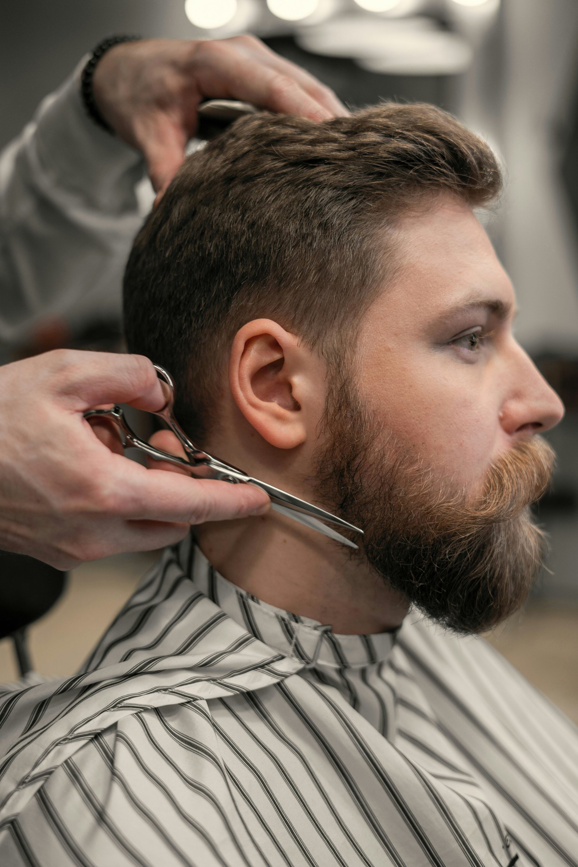 A barber trimming a man's beard with scissors in a salon